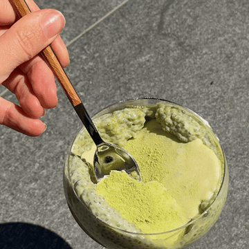 Ein Glas mit Matcha Chia Pudding und einer Schokoladendecke, in die mit einem Löffel gestochen wird. 